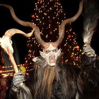 A frightening Krampus with horns and a long mane stands amidst a crowd of people. In the background, Christmas lights shine and a large Christmas tree is illuminated. | © Verein Salzburger Christkindlmarkt