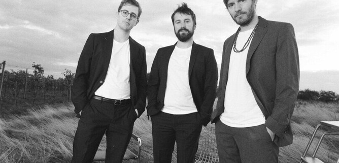 Three men in suits stand in an open field. They appear self-confident and are photographed in a modern, artistic black-and-white style. | © Julian Hagen