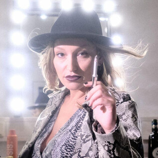 Eine Frau mit einem schwarzen Hut und einem Schlangenmuster-Jacket hält einen Stift in der Hand. Der Hintergrund ist mit Spiegellichtern beleuchtet. | © Christopher Glanzl