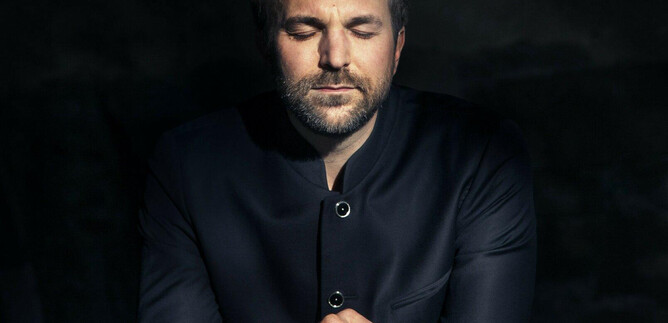 A man with closed eyes sits calmly in a dark environment. He is wearing a black jacket and has a thoughtful demeanor. | © Felix Broede