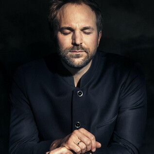 A man with closed eyes sits calmly in a dark environment. He is wearing a black jacket and has a thoughtful demeanor. | © Felix Broede