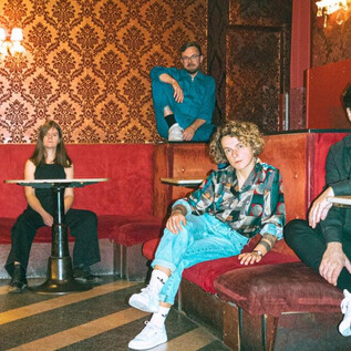 A group of four people is sitting and standing in a stylishly decorated room with red decor. The atmosphere is relaxed and creative. | © Julia Freytag