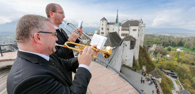 Two musicians are playing trumpets with an impressive castle in the background. The scene showcases a blend of culture and history against a picturesque landscape. | © www.salzburg-burgen.at