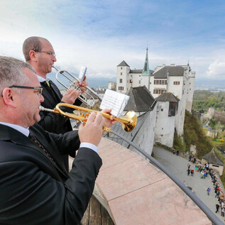 Zwei Musiker spielen auf Trompeten mit einer beeindruckenden Burg im Hintergrund. Die Szenerie zeigt eine Mischung aus Kultur und Geschichte vor einer malerischen Landschaft. | © www.salzburg-burgen.at