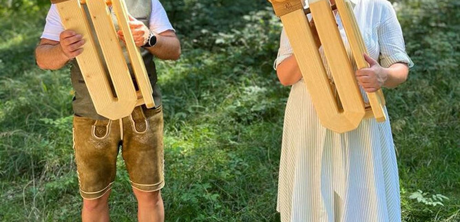 D dvě osoby stojí v lese a hrají na velkých dřevěných dechových nástrojích. Muž má na sobě tradiční kroj, zatímco žena je oblečena ve světlých šatech. | © Salzburger Freilichtmuseum