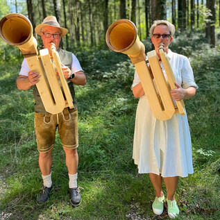 Zwei Personen stehen im Wald und spielen auf großen Holzblasinstrumenten. Der Mann trägt traditionelle Tracht, während die Frau in einem hellen Kleid gekleidet ist. | © Salzburger Freilichtmuseum