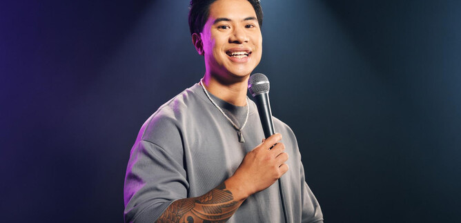 A young man with a microphone smiles at the camera. He is wearing a gray sweatshirt and is standing in front of a dark, illuminated background. | © Marvin Ruppert