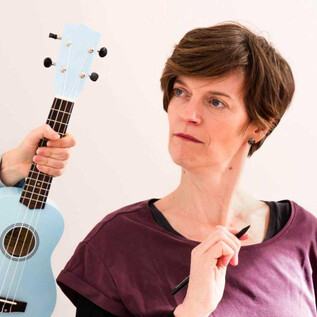 Eine Frau schaut nachdenklich, während jemand im Hintergrund eine hellblaue Ukulele hält. Sie scheint einen Stift in der Hand zu haben und ist in einem kreativen Moment. | © Klemens Dellacher