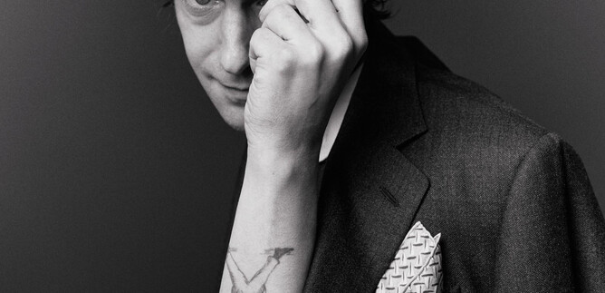 Ein junger Mann trägt einen Hut und ein elegantes Sakko. Er blickt spielerisch in die Kamera und zeigt ein Tattoo auf seinem Arm. | © Susanne Hassler-Smith