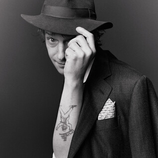 A young man is wearing a hat and an elegant jacket. He playfully looks at the camera and shows a tattoo on his arm. | © Susanne Hassler-Smith