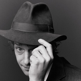 Ein Mann trägt einen Hut und schaut lächelnd zur Seite. Die Aufnahme ist in Schwarz-Weiß gehalten. | © Susanne Hassler-Smith