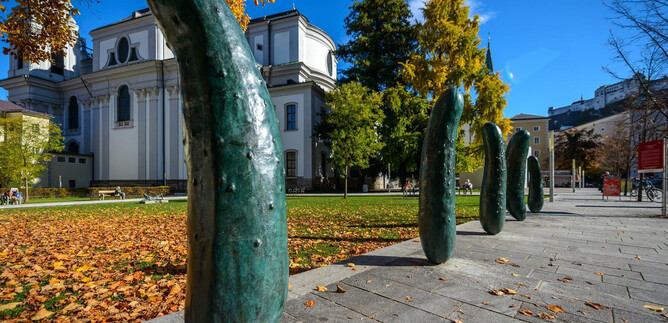 An interesting series of sculptures made of green bronze forms stands in a square. In the background, you can see trees and a historic building. | © TSG