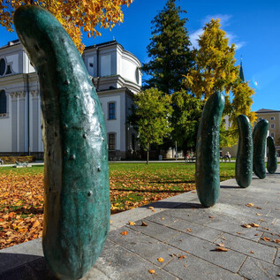 An interesting series of sculptures made of green bronze forms stands in a square. In the background, you can see trees and a historic building. | © TSG