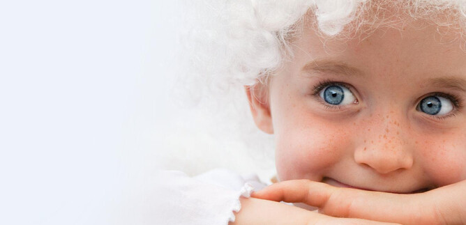 Uśmiechnięte dziecko z niebieskimi oczami i kręconymi włosami, które podpiera się na rękach. Tło jest jasne i neutralne. | © Stiegl-Brauwelt