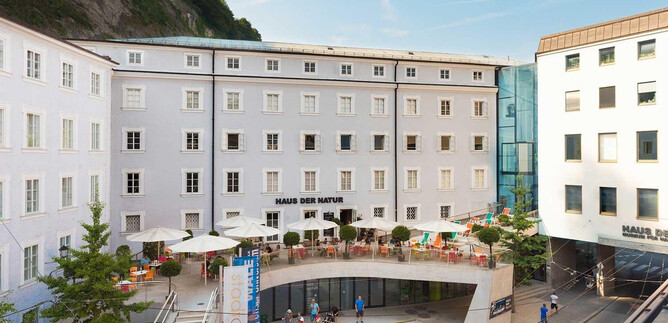 A modern building with a bright facade and glass accents. In front of the building, there are terraces with seating and plants. | © HdN_Simmerstatter