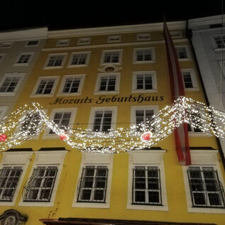 Un edificio storico, noto come la casa natale di Mozart, brilla di notte. La facciata è decorata con luci festive e decorazioni. | © ISM