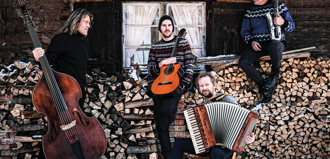 A musical group stands in front of a woodpile in a wintery environment. The musicians play various instruments and wear warm sweaters. | © Quadro Nuevo