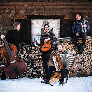 Eine musikalische Gruppe steht vor einem Holzstapel in einer winterlichen Umgebung. Die Musiker spielen verschiedene Instrumente und tragen warme Pullover. | © Quadro Nuevo
