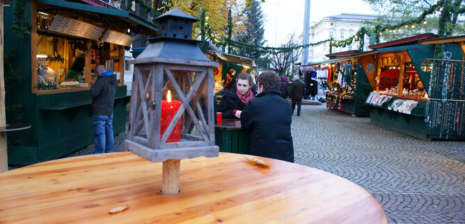 Un mercado de Navidad acogedor con puestos decorados festivamente y personas conversando. Una linterna con luz roja está en primer plano sobre una mesa de madera.