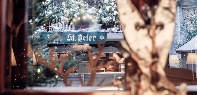 Ein Blick aus einem Fenster auf eine festlich dekorierte Umgebung mit Schnee und einem Schild, das "St. Peter" zeigt. Im Vordergrund sind dekorative Elemente zu sehen, die eine gemütliche Atmosphäre schaffen. | © St. Peter Stiftskulinarium