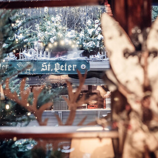 Ein Blick aus einem Fenster auf eine festlich dekorierte Umgebung mit Schnee und einem Schild, das "St. Peter" zeigt. Im Vordergrund sind dekorative Elemente zu sehen, die eine gemütliche Atmosphäre schaffen. | © St. Peter Stiftskulinarium