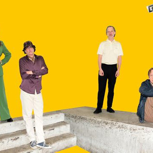 A group of four people stands on a concrete platform in front of a yellow background. They are dressed differently and posing confidently. | © Stefan Braunbarth