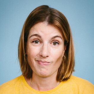 A woman with shoulder-length hair is wearing a yellow T-shirt. She has a neutral facial expression against a blue background. | © Apollonia Theresa Bitzan