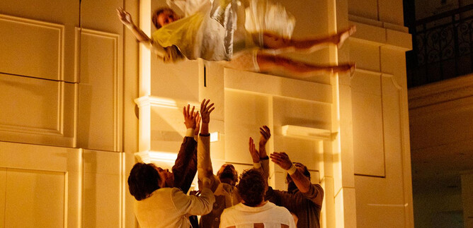 Eine Person schwebt in einem hellen Raum, während mehrere Leute sie auffangen wollen. Die Szene vermittelt eine dramatische und spannende Atmosphäre. | © Le Bon Marché_Rive Gauche