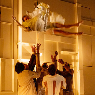 Eine Person schwebt in einem hellen Raum, während mehrere Leute sie auffangen wollen. Die Szene vermittelt eine dramatische und spannende Atmosphäre. | © Le Bon Marché_Rive Gauche