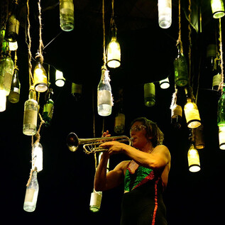 Ein Musiker spielt Trompete unter schwebenden Flaschenlichtern. Die Atmosphäre ist kreativ und stimmungsvoll. | © Cecile Gerbe Servettaz
