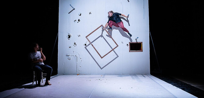 A dramatic scene with an acrobat hanging on a wall. In the foreground, a person sits on a chair and watches the performance. | © Cosmin Cirstea
