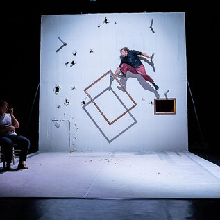 Eine dramatische Szene mit einem Akrobaten, der an einer Wand hängt. Im Vordergrund sitzt eine Person auf einem Stuhl und beobachtet die Darbietung. | © Cosmin Cirstea