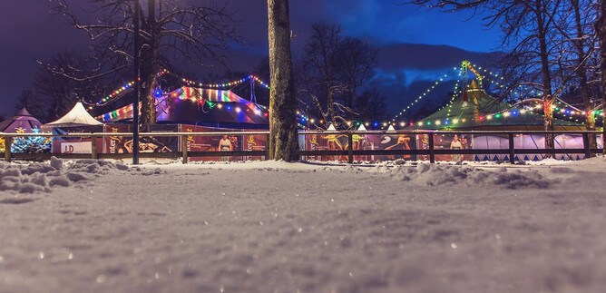 A snowy winter landscape with colorful lights and festive decorations. In the background, festive stalls and trees can be seen. | © Winterfest / Eva trifft 