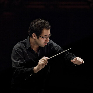 A conductor stands on stage and conducts with a baton. Behind him, a dark backdrop is visible. | © Pia Clodi