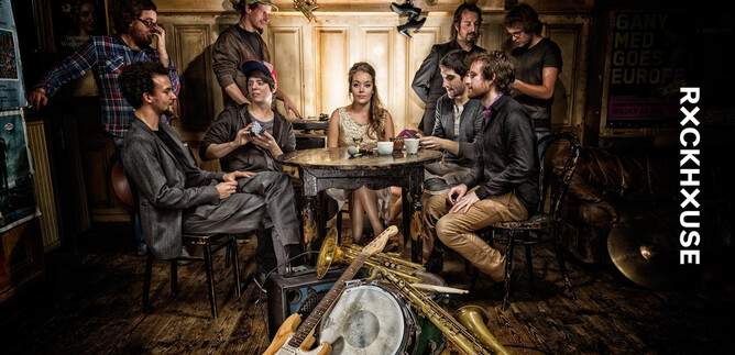 A group of people is sitting at a table in a cozy room. Instruments like trumpets and a drum set are lying on the floor. | © Rockhouse Salzburg