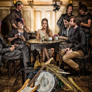 Eine Gruppe von Menschen sitzt an einem Tisch in einem gemütlichen Raum. Auf dem Boden liegen Instrumente wie Trompeten und ein Schlagzeug. | © Rockhouse Salzburg