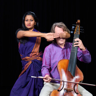 A man is playing a musical instrument while a woman behind him covers his eyes with one hand. Both are wearing traditional clothing. | © Raphael Mittendorfer