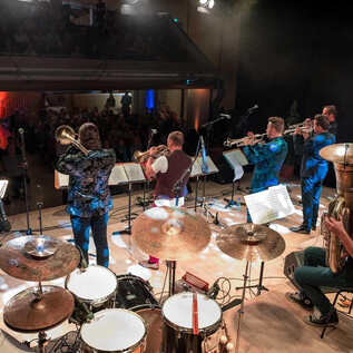 Eine Band spielt auf einer Bühne mit verschiedenen Instrumenten. Die Musiker stehen in einem festlichen Raum mit einer begeisterten Menge im Hintergrund. | © bodenst@ndig