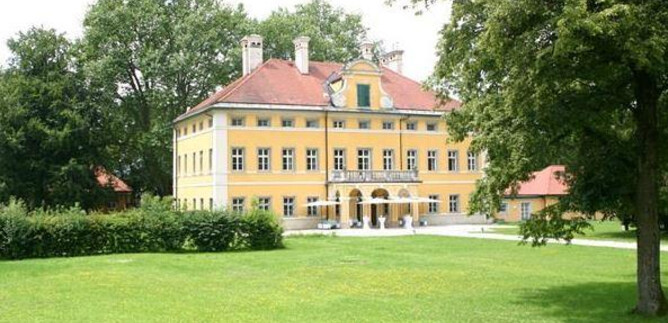 Velká žlutá budova s červenou střechou stojí v zelené krajině. Kolem budovy jsou stromy a udržovaná tráva. | © Christian Schneider