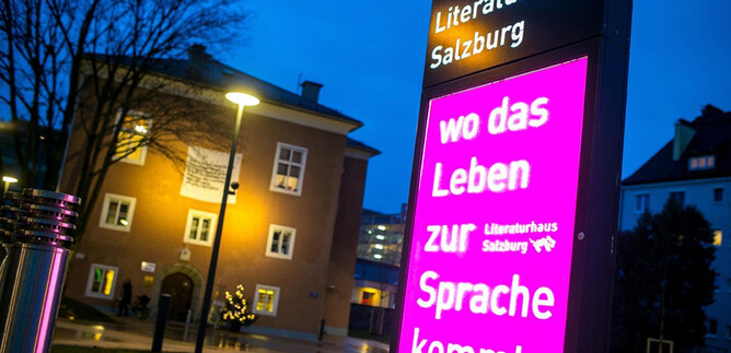 Ein beleuchtetes Schild vor dem Literaturhaus Salzburg zeigt den Slogan „wo das Leben zur Sprache kommt“. Im Hintergrund ist ein modernes Gebäude und Bäume zu sehen. | © Literaturhaus Salzburg