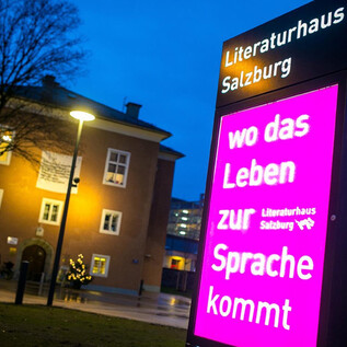 Una señal iluminada frente a la Casa de la Literatura de Salzburgo muestra el lema "donde la vida se expresa". Al fondo se puede ver un edificio moderno y árboles. | © Literaturhaus Salzburg
