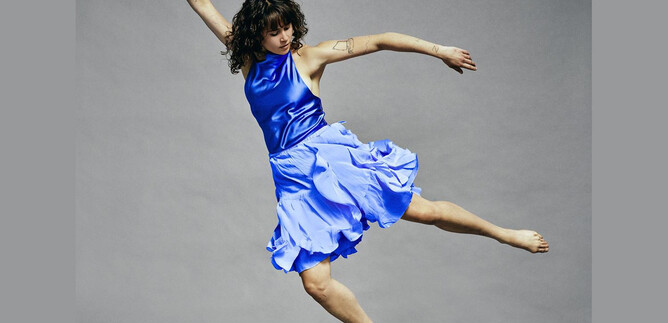 A dancer in a blue dress jumps dynamically through the air. The background is gray and neutral. | © Chris Rogl