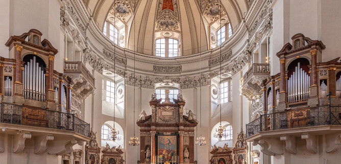 A magnificent church with a high dome and elaborate ceiling frescoes. Benches and organs flank the altar in the center. | © Salzburger Dom / Eva trifft. Fotografie