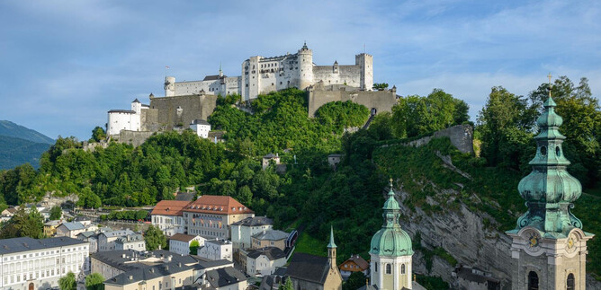Un castello impressionante su una collina, circondato da verde natura. In primo piano sono visibili i tetti di una città con edifici storici. | © Tourismus Salzburg / G. Breitegger