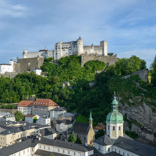 Impozantní hrad na kopci, obklopený zelenou přírodou. V popředí jsou vidět střechy města s historickými budovami. | © Tourismus Salzburg / G. Breitegger