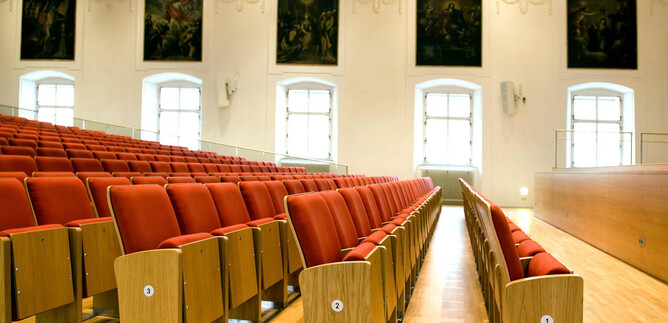A large hall with red seats and wooden paneling. Paintings hang on the walls, and large windows provide plenty of daylight. | © Universität Salzburg, Luigi Caputo