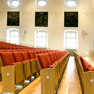A large hall with red seats and wooden paneling. Paintings hang on the walls, and large windows provide plenty of daylight. | © Universität Salzburg, Luigi Caputo
