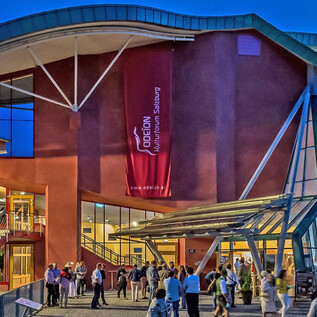 A modern building with large window fronts and a red banner. People are gathering in front of it and enjoying the evening atmosphere. | © Albert Moser