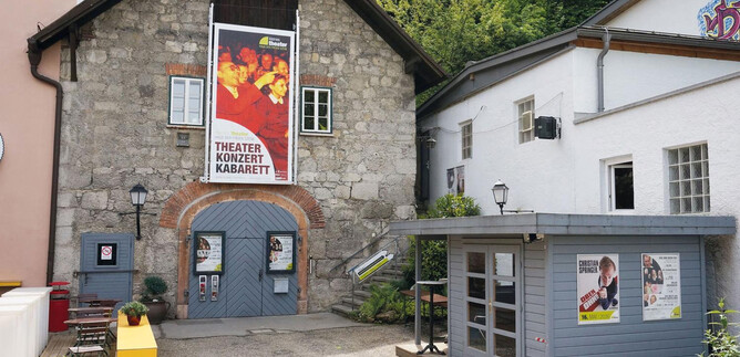 Ein charmantes Theatergebäude mit einer großen Plakatwand, das kulturelle Veranstaltungen anbietet. Nebenan befindet sich ein kleiner Kiosk mit bunten Anzeigen. | © Kleines Theater