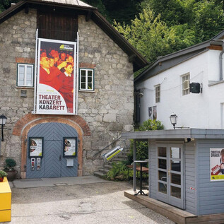 Un charmant bâtiment de théâtre avec un grand panneau d'affichage, qui propose des événements culturels. À côté se trouve un petit kiosque avec des annonces colorées. | © Kleines Theater